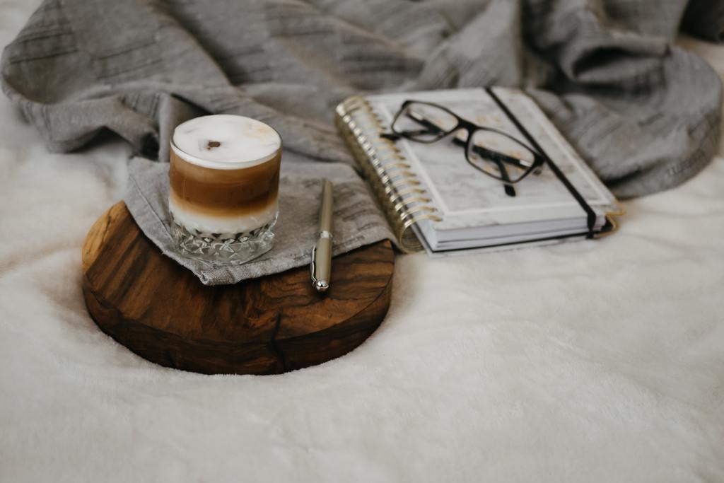 image of coffee placed on a wooden coaster on a bed with a notebook, pen and reading glasses beside it.